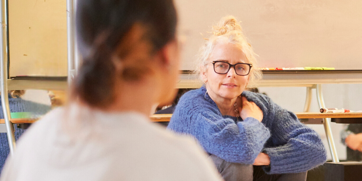 Blonde-haired facilitator wearing glasses and a blue sweater listening to youth during a session Blonde-haired facilitator wearing glasses and a blue sweater listening to youth during a session